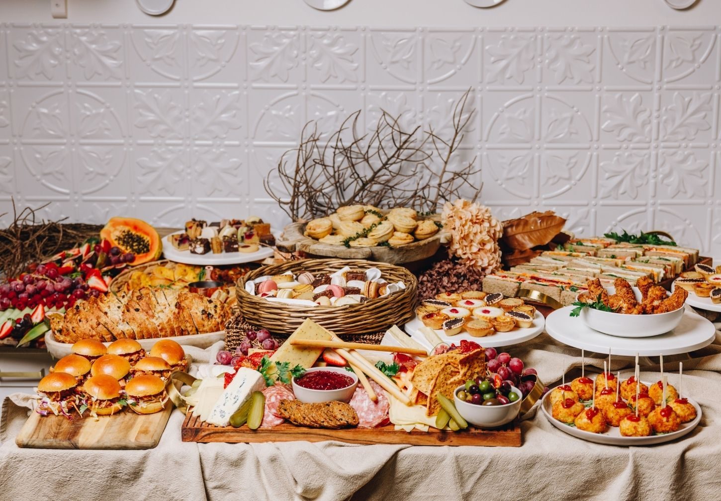 Catering spread with sandwiches, sliders, pastries, fruit, cheese, cured meats and assorted finger food.